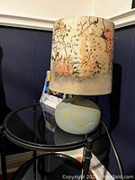 Side view of the lamp showing painted pastel base and floral decoupage paper shade on a reflective black glass table against a dark paneled wall.