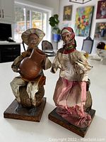 The photo shows two Mexican papier mâché figurines placed on a table indoors near a window. One figurine is a man seated with a jug, the other is a woman standing wearing a headscarf.