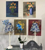 Full view of the two lamps displayed against a wall with floral paintings. Lamps have white bases and blue floral decoupaged shades.