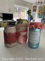 Photo of three decorated jars with various designs placed on a wooden surface in front of a kitchen window.