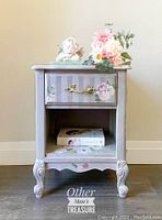 Front view of the small French Provincial night table showing one drawer with ornate gold-tone handle, floral hand-painted design on drawer and frame, purple vertical stripes on drawer, curved legs, and an open lower shelf with books inside.