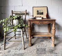 Photo showing the entire side table made of wood with detailed carved legs and bottom shelf, with staged items including an old chair, a plant, and framed art behind.