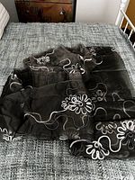 Three black sheer curtain panels with white floral and swirl embroidery laid on a bed.