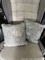 Photo showing two light blue square throw cushions in toile fabric with detailed pastoral patterns placed on a chair.
