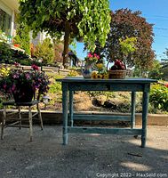 Wooden work table with distressed blue-green paint finish, placed outdoors with plants in the background.