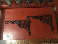 Two black, heavy iron decorative brackets for hanging plants or chimes laid out on a red-painted wooden surface. Each bracket features scrollwork and floral motif design elements with a curved hook at one end.