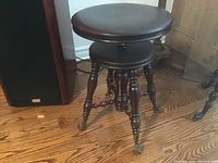 Side view of antique piano stool showing round padded seat, turned wooden legs, and castors for mobility.