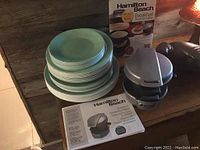 Stacked light blue-green Correlle dishes (plates and bowls) next to boxed Hamilton Beach Breakfast Sandwich Maker with unit outside box on rustic wooden surface.