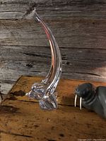 Crystal dolphin sculpture with clear abstract form and curved elongated shape on wooden surface with walrus figurine nearby.