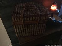 Wicker picnic basket on wooden table under low light showing woven texture and two handles closed.