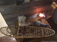 Photo showing wooden snowshoes with woven webbing and leather bindings alongside tan leather snowshoe boots and an antique oil lamp on a wooden table.