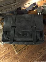 Front view of the grey/blue Roots messenger bag with two vertical straps and 'BOURNE LEGACY' embossed on leather patch.