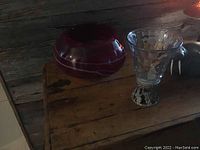 Two items on a wooden surface against a rustic wood backdrop: a round cranberry red glass bowl and a pewter tealight candle holder with a textured base.