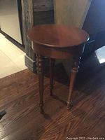 Small round wooden side table with three turned legs, shown on wooden floor next to rustic wooden panel.