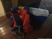Photo showing three life jackets in different colors and one large blue cooler with white lid and cup holders