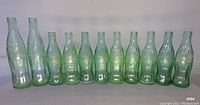 Photo showing 11 green tinted Coca Cola glass bottles side by side on a white surface, various sizes including mostly 6 oz. and 2 larger 10 oz. bottles.