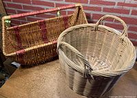 Two large vintage wicker baskets shown together, one round and one rectangular, on a wooden surface against a brick wall.