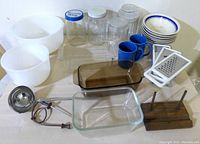 Full top-down view of all items in lot showing mixing bowls, glass loaf pans, jars, mugs, grater, ladle, wooden stand, plates