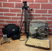 Wide shot showing all items in the lot including camera bags, two tripods, vintage projector with power cord, and accessories arranged on table against brick wall.