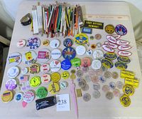 Photo showing full assortment of pinback buttons featuring various slogans and designs, advertising pencils arranged in rows, wooden nickels with promotional printing, and small collectible items on a table.