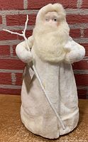 Full figure of Department 56 Snow Angels Father Christmas in white coat holding white branch staff against red brick background