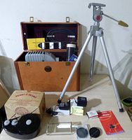 View of the Kodaslide Master Model slide projector inside brown carrying case, power adapter, lens caps, slide holders, tripod extended with handle and adjustment knob.