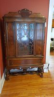 Full view of antique wooden china cabinet standing on turned legs with drawer and glass door.