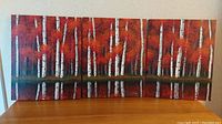 Full view of three connected canvas paintings depicting birch trees with red and orange foliage on a wooden surface.