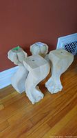 Four light wooden claw foot style table legs arranged on wooden floor, showing carved ball and claw design and square top sections.