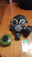 Front view of the black rubber gas mask showing the eye lenses, breathing apparatus, and green metal canister beside it on the wooden surface.