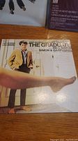 Album cover of The Graduate soundtrack showing a man in a suit and a leg extended in stockings; includes text naming Simon & Garfunkel.