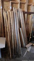 Photo showing bundle of wooden stakes leaning against a wall in a storage area with plywood paneling, stakes vary slightly in color and some have visible wood knots.
