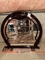 Front view of a round mirror with black painted wooden composite frame, decorative trim and flat base, placed on carpet with various storage boxes in the background.