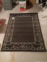 Grey rectangular carpet with cream scroll decorative border, laid flat on floor with some surrounding household items.