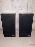 Front view of a pair of Accu-Tech AR-4L speakers with black fabric grilles and wood veneer side panels.