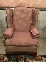 Front view of vintage wingback chair showing dusty rose fabric with floral pattern, padded arms, and wooden legs.