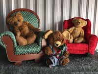Three fully jointed teddy bears posed together with two miniature upholstered chairs, on a dark surface against a metal background