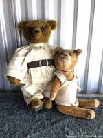 Two bears sitting side by side on a dark surface with metal background, one wearing safari outfit and the other in white sleeveless shirt with glasses.