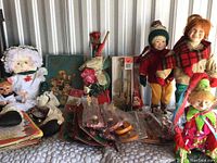 Photo showing an assortment of holiday dolls, figurines, books, and decorations arranged on a table including fabric dolls with winter clothing and a Santa tree-topper
