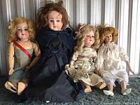 Four dolls seated side by side showing different dress colors and doll types (porcelain and wooden).