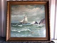 Front view of framed maritime painting showing sailboat, ocean waves, rocky cliffs, and cloudy sky with artist signature visible.