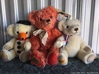 Three Merrythought teddy bears sitting side by side: sun-bleached red, white with ribbon, and white dressed as snowman.