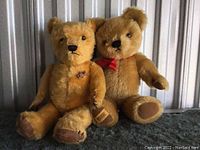 Two Merrythought teddy bears, one with a red ribbon bow and other with a small teddy pin, sitting together on a textured surface.
