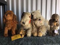 Front view of four mohair dog plush toys, varying in size and color, with glass eyes displayed on a dark surface against a metal backdrop.