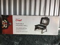 Close-up photo of the original box showing Master Chef Portable Propane Grill with 10,000 BTUs, porcelain-coated steel grill surface and cook area.