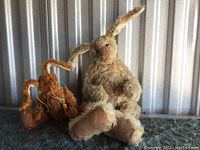 Two mohair rabbit dolls sitting on a dark surface against a corrugated metal background. One cream-colored, larger, fully jointed, about 15.5 inches. One smaller brown with a carrot pendant around its neck.