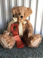 Full view of the golden teddy bear seated against a corrugated metal background. The bear is about 20 inches tall with cream ribbon around neck and a large orange award ribbon attached.