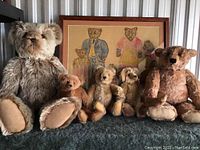 Photo showing full view of five mohair teddy bears of varying sizes arranged in front of framed needlepoint bear family art, on a dark blue textured surface.