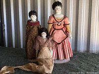 Three vintage porcelain dolls standing and sitting on a surface, showing full-body views of each doll's clothing and heads.
