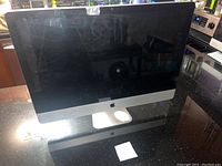 Front view of 27-inch 2010 Apple iMac with black screen off, on a black granite surface, showing aluminum frame and Apple logo.